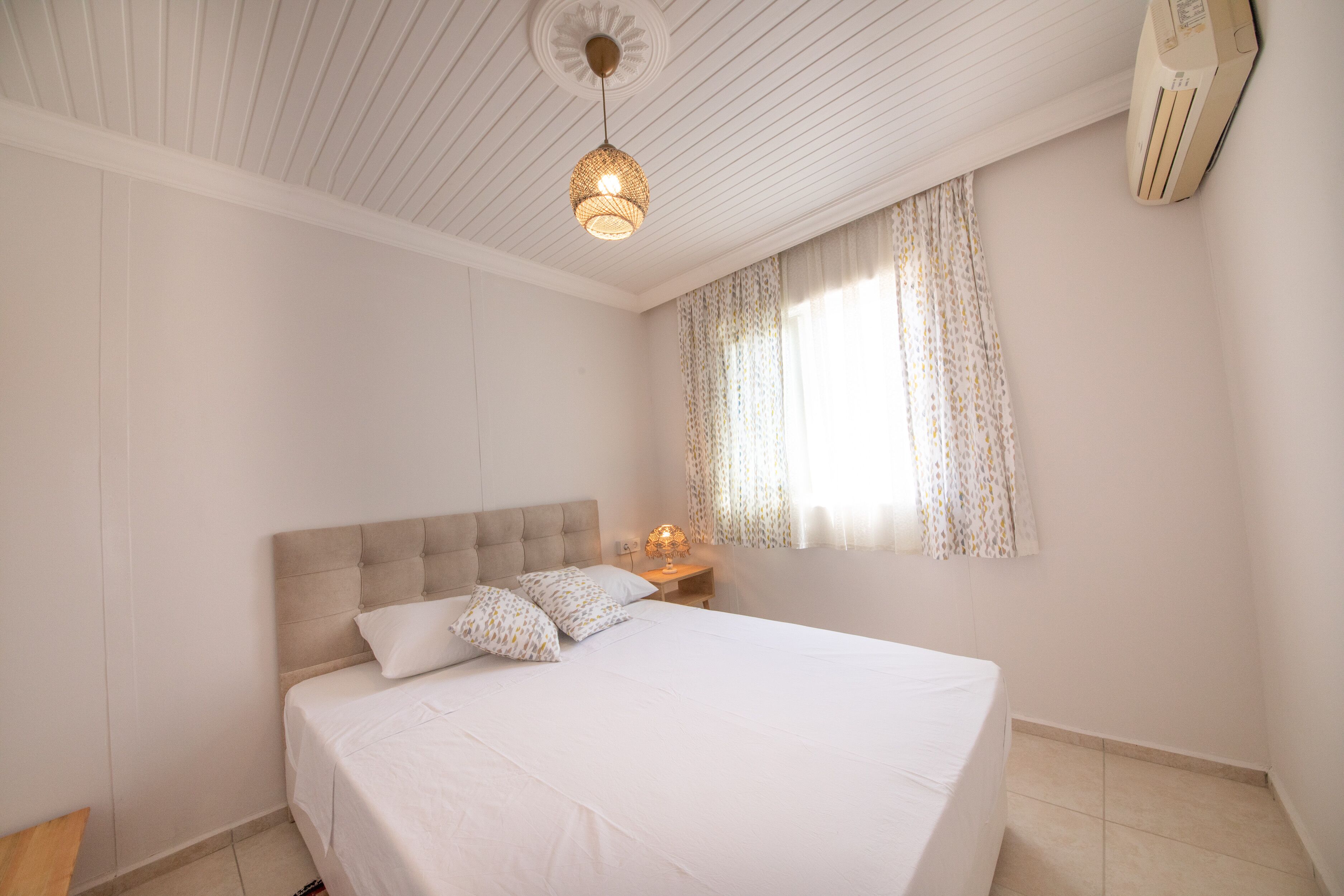 A bedroom with a bed, a bedside table with a lamp, a window with curtains, and an air conditioning unit.