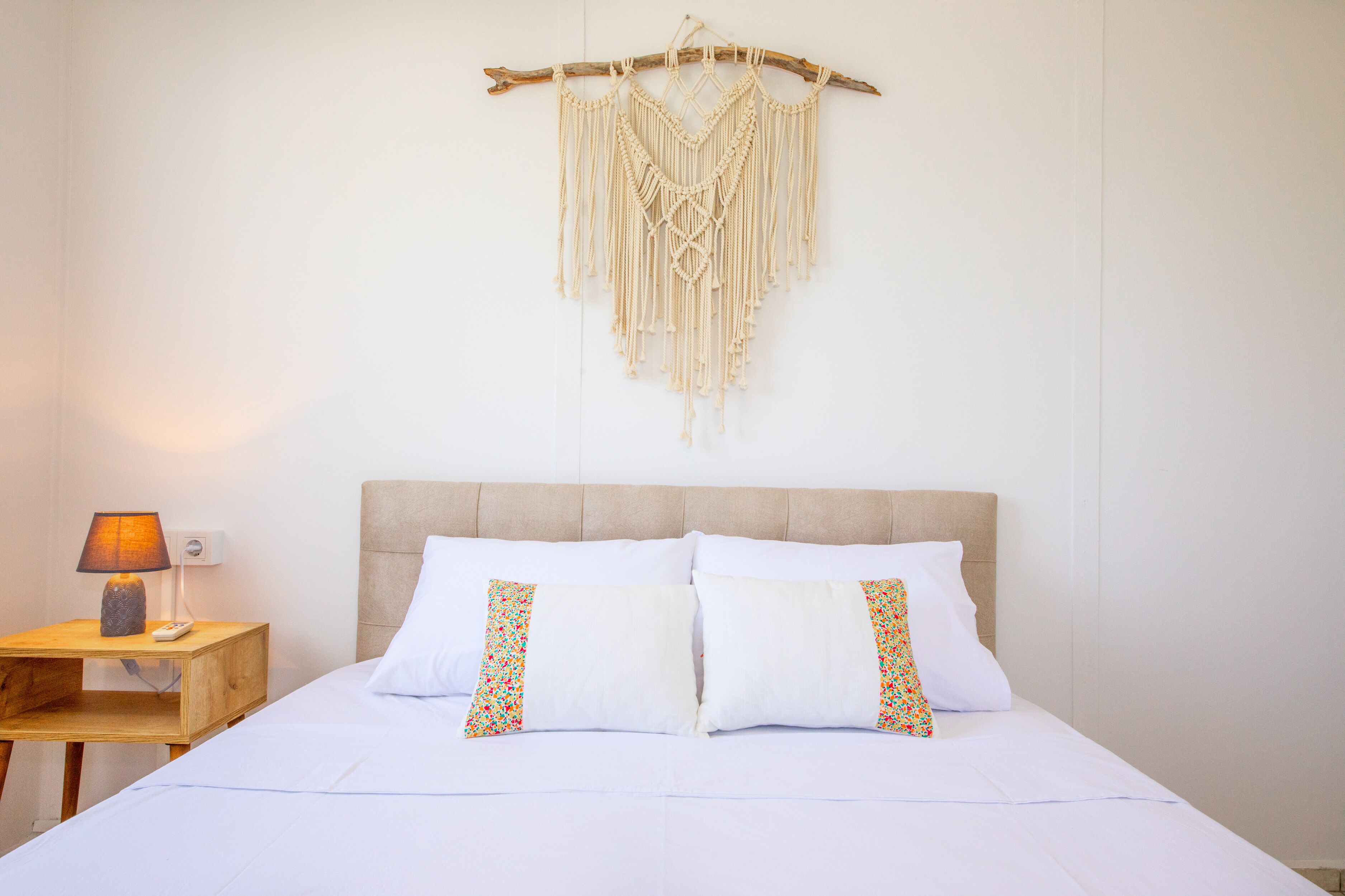 Una cama bien hecha con dos almohadas, una mesita de noche de madera con una lámpara y un colgador de macramé en la pared.