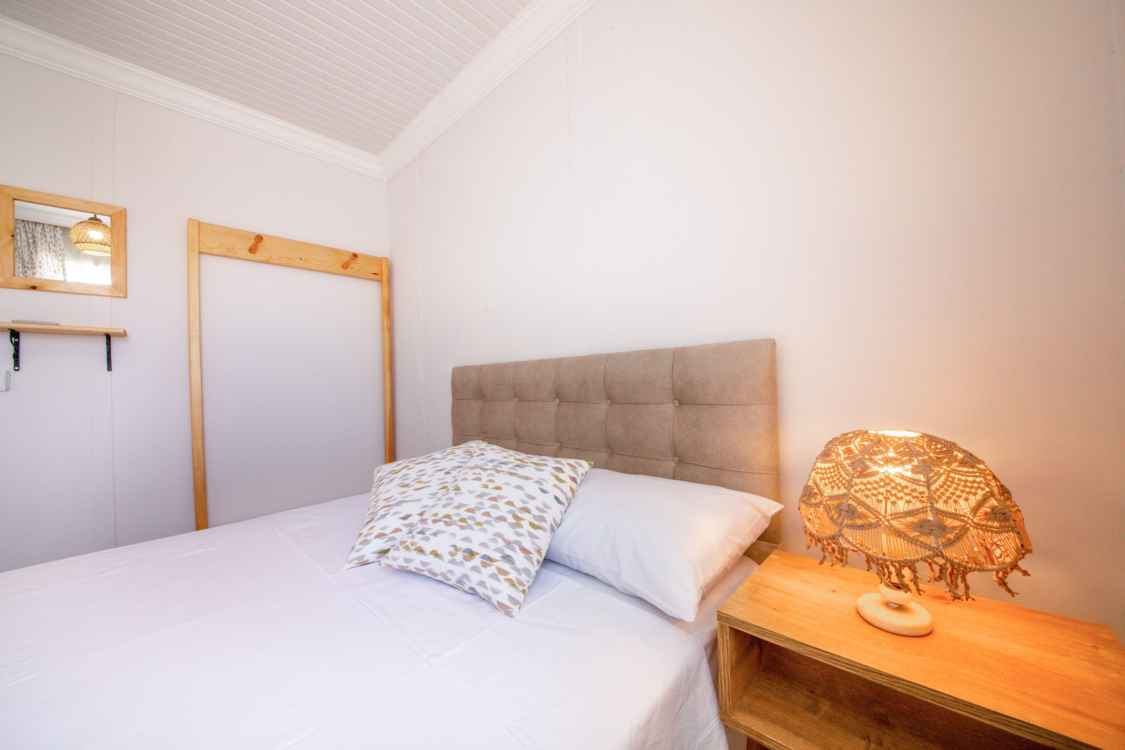 Uma cama bem arrumada com travesseiro estampado, cabeceira de madeira e abajur com cúpula trançada.