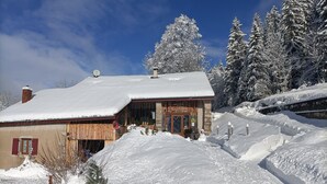 A snow-covered building with a wooden facade, a visible entrance, and a snow-covered landscape with trees. - Gîte la grange de l'errance (Giron)
