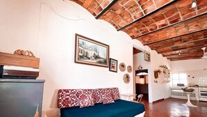 A room with a bed, a small table, a kitchen area, and a brick ceiling.