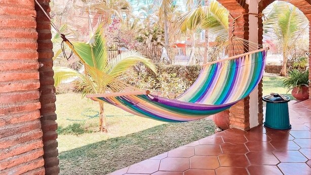A hammock hanging between brick arches in a covered outdoor area with a view of palm trees and a garden.