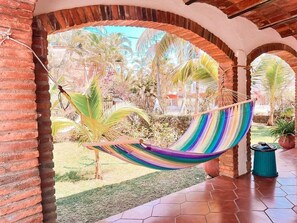 Un hamac est suspendu entre des arcades en briques dans un espace extérieur abrité, offrant une vue sur des palmiers et un jardin.