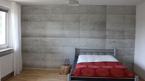 A bedroom with a concrete wall, a bed with a red patterned bedspread, a small wooden stool, and a window with white curtains.