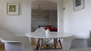 A modern dining area with a round white table, two white chairs, and a vase with red flowers. There are framed artworks on the walls and a fireplace in the background.