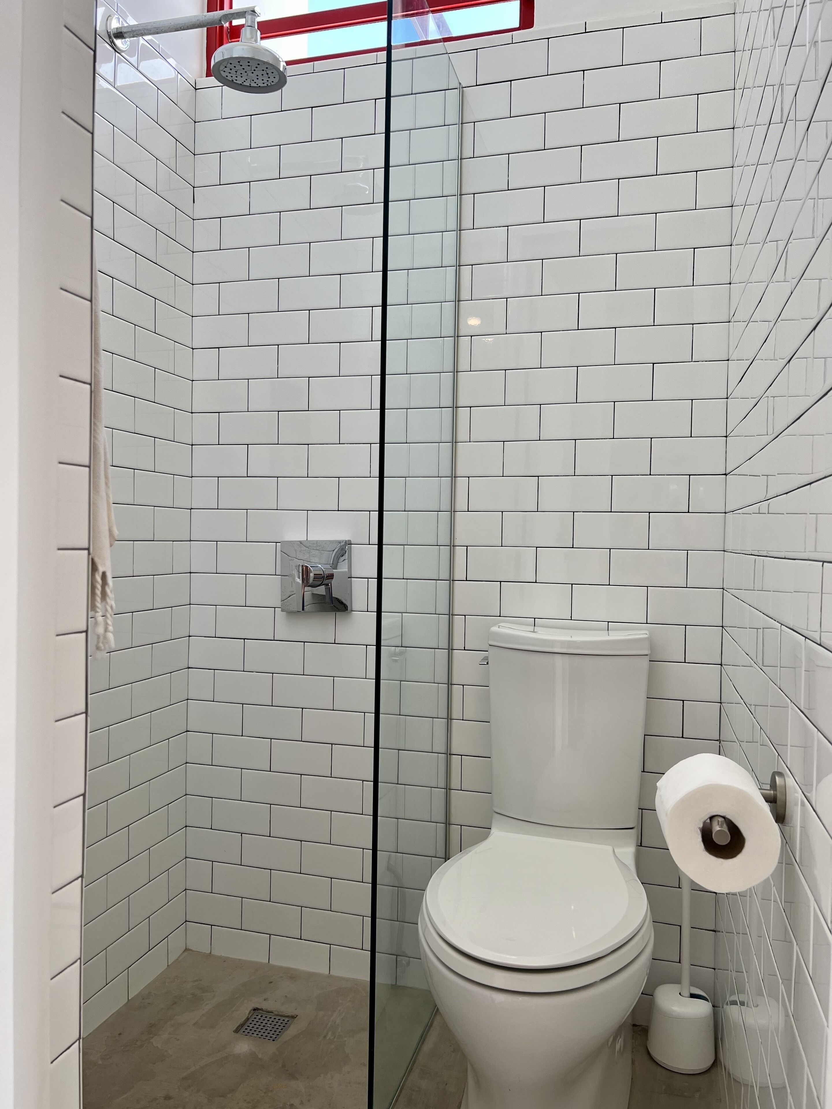 Un baño con azulejos blancos tipo metro, cabina de ducha de vidrio, inodoro y dispensador de toallas de papel.