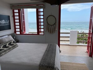 Une chambre avec un lit, un attrape-rêves et une vue sur l’océan, visible à travers des portes rouges ouvertes.