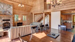 L’intérieur d’une cabane en rondins présente une cheminée en pierre, un coin repas avec une table et des chaises en bois, un espace cuisine et un escalier menant à un niveau supérieur.