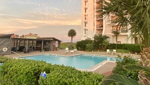 Une piscine entourée de verdure, un immeuble imposant doté de balcons et un ciel dégagé.