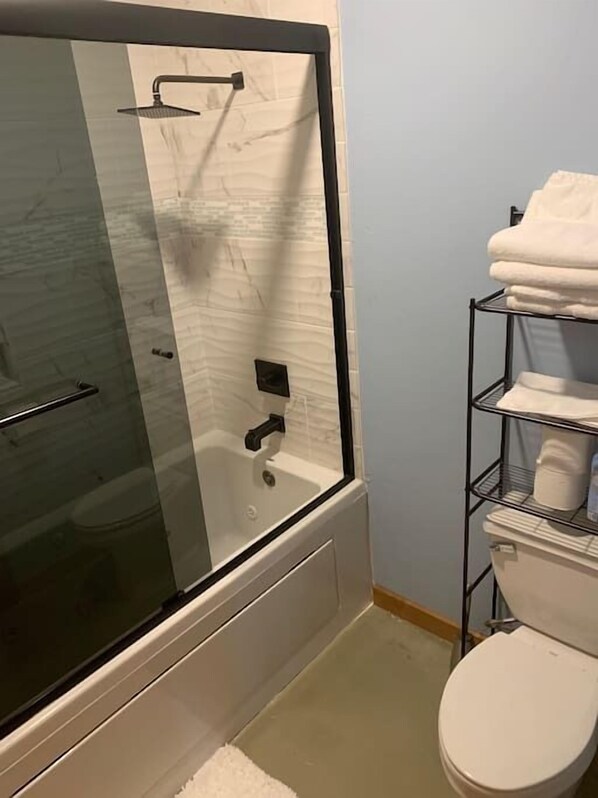 A bathroom with a glass-enclosed shower, a white toilet, and a metal shelf with towels. - Jeremys Mountain Chalet @jeremysmountainchalet (Anchorage)