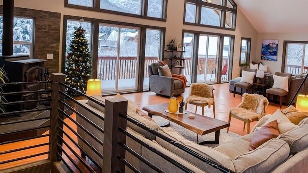 A spacious living area with a high ceiling, large windows, a fireplace, and a Christmas tree. - Jeremys Mountain Chalet @jeremysmountainchalet (Anchorage)