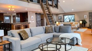 A spacious living area with a large gray sectional sofa, a glass coffee table, and a wooden staircase leading to an upper level. - Jeremys Mountain Chalet @jeremysmountainchalet (Anchorage)