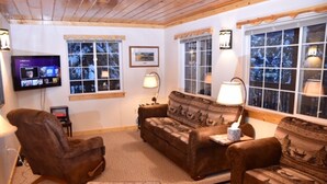 A cozy living room with a brown leather sofa, a brown armchair, and a patterned ottoman. There are two windows with curtains, a television on the wall, and a framed picture above the sofa. - Eagle's Landing, is located in Island Park, ID, 35 miles from West Yellowstone (Island Park)