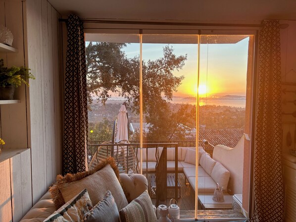 A balcony with a sofa and a table, overlooking a scenic view of trees and a sunset.