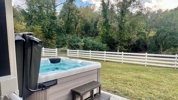 A hot tub with a wooden bench and a small table, surrounded by a white fence and lush greenery.