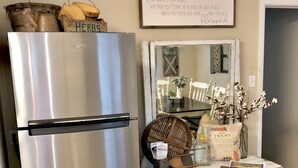 A stainless steel refrigerator, a white wooden chest of drawers, and a mirror reflecting a dining table with chairs.