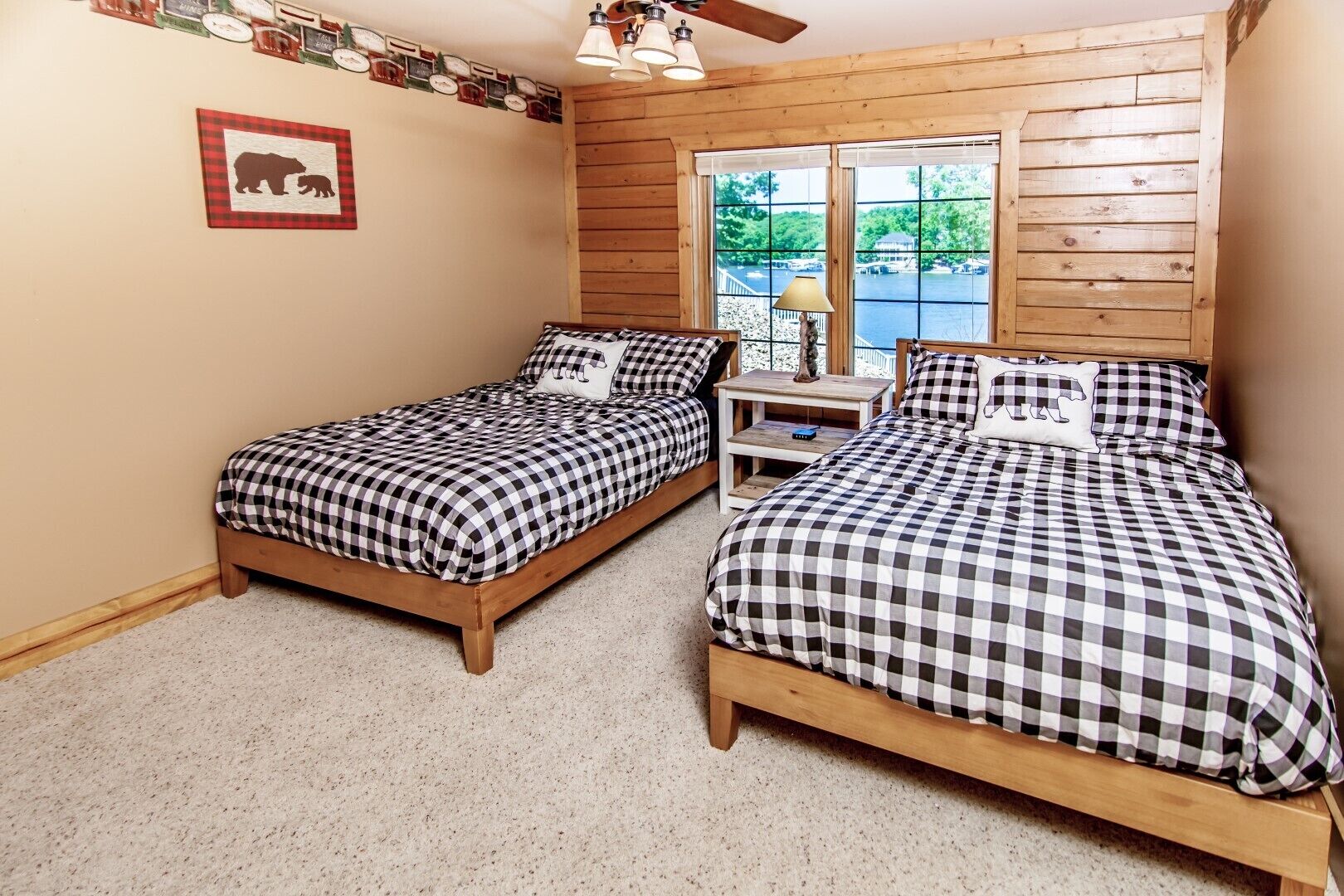 Una habitación con dos camas de madera, ropa de cama a cuadros, una mesita, una lámpara y una ventana con vista a un muelle y barcos.