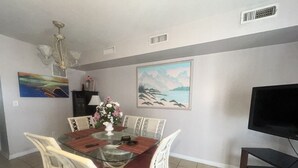 A dining area with a glass table surrounded by white wicker chairs, a vase with flowers, and a painting of a beach scene.