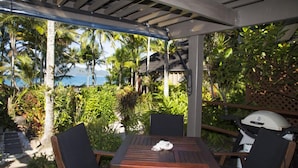 A covered outdoor seating area with a table and chairs, surrounded by lush greenery and a view of the ocean. - Stay at ‘Banfield 3' (Wongaling Beach)