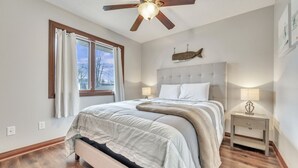 Un dormitorio con cama, ventilador de techo, mesita de noche, lámpara, ventana con cortinas y decoración de peces en la pared.