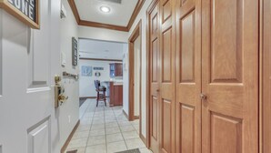 Un pasillo con una puerta de madera, suelo de baldosas y una pequeña cocina al fondo.