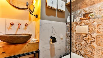Casa de banho com toucador de madeira, lavatório em forma de taça, espelho, porta-toalhas e azulejos estampados.
