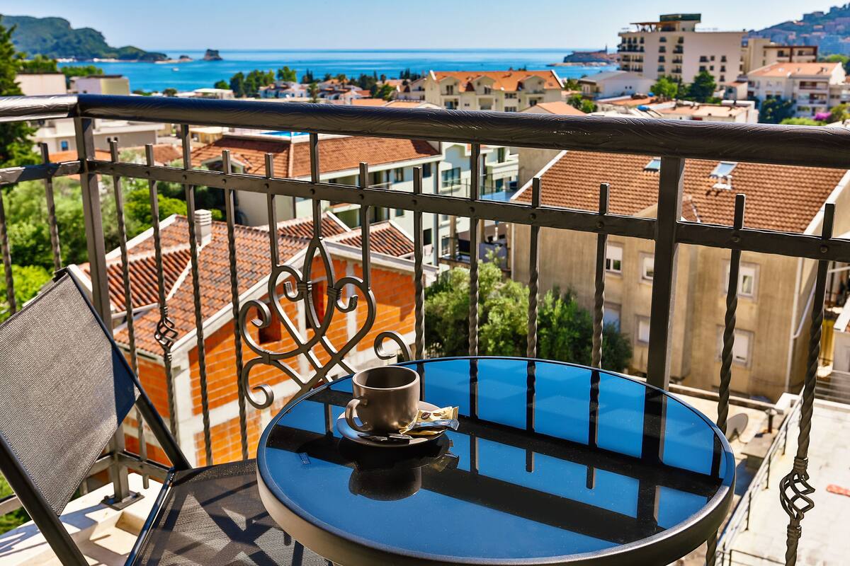 A balcony with a table and chair, overlooking a coastal town with terracotta rooftops and buildings.