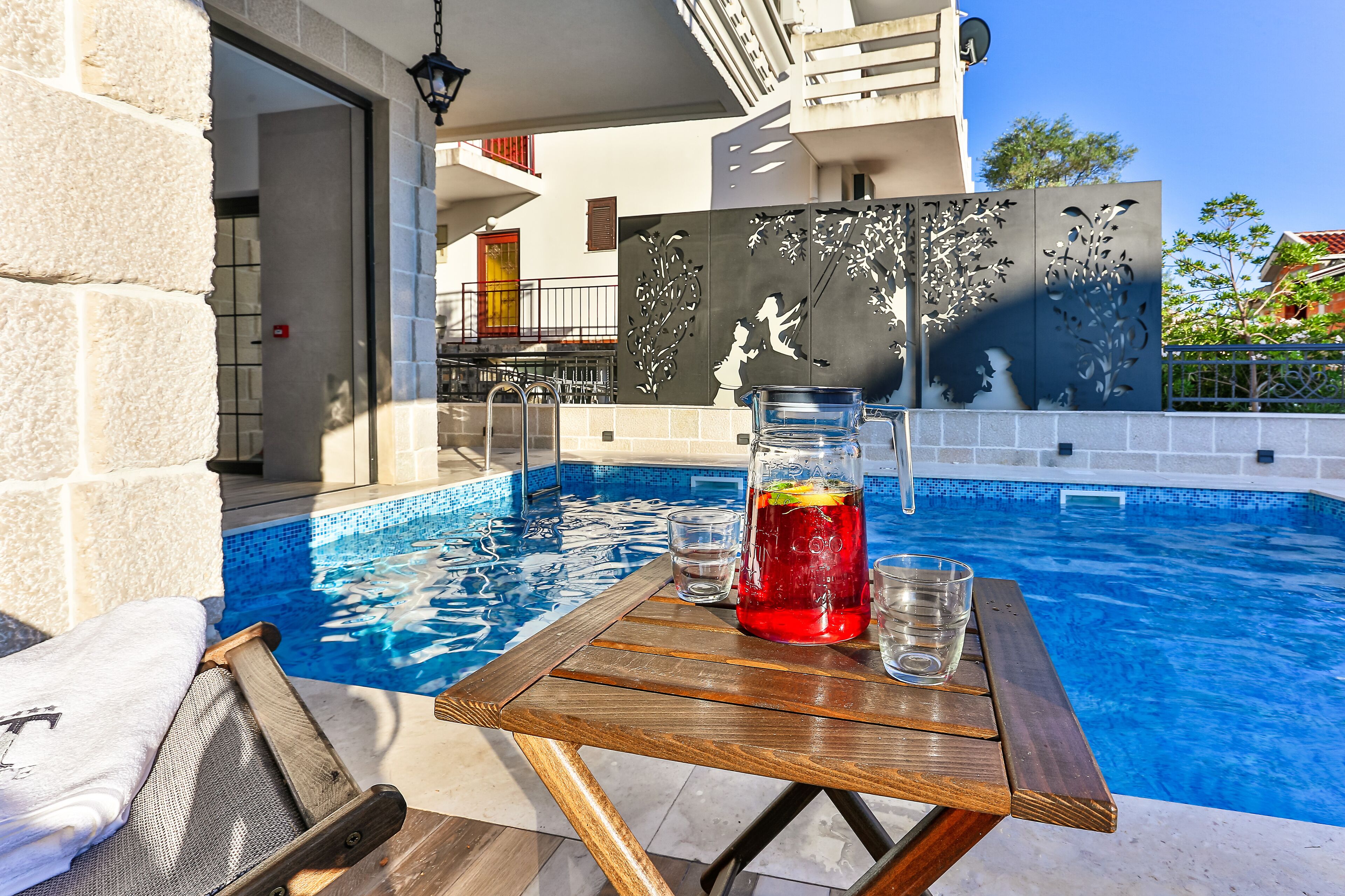 A poolside area with a wooden table, a pitcher of red drink, and clear glasses.
