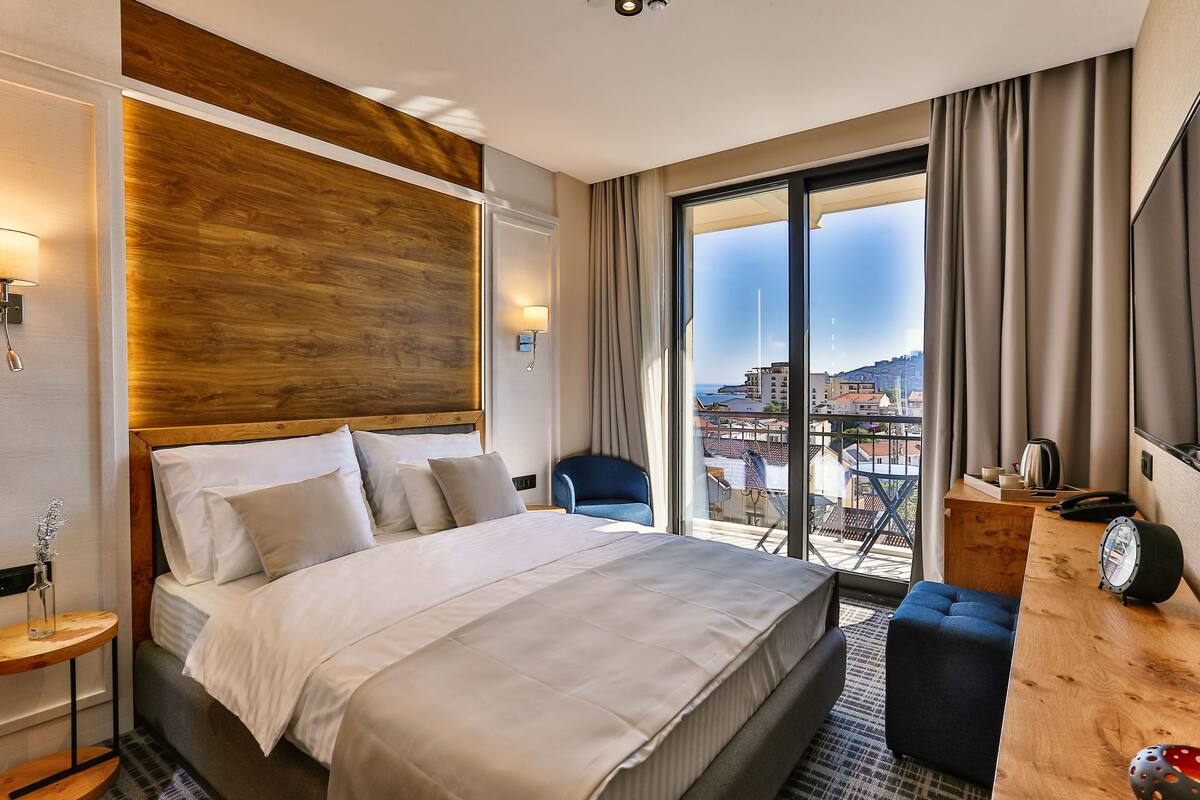 A modern hotel room with a large bed, a wooden headboard, a desk with a telephone, and a view of the city through large windows.