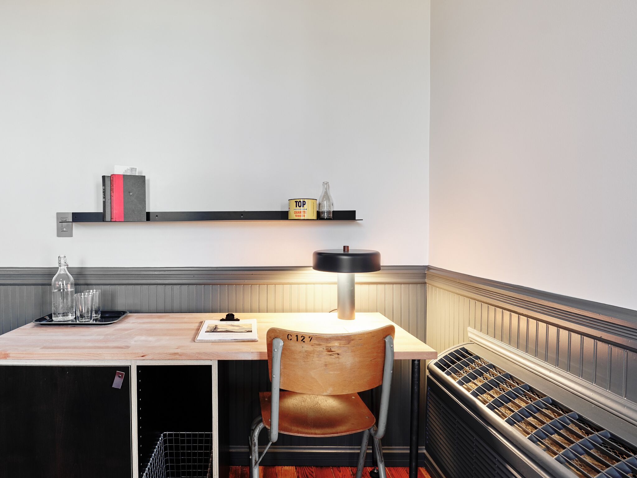 A corner workspace with a wooden table, a metal chair, a lamp, a shelf with books and a cigar tin, and a radiator.
