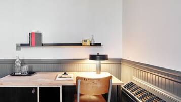 A corner workspace with a wooden table, a metal chair, a lamp, a shelf with books and a cigar tin, and a radiator.