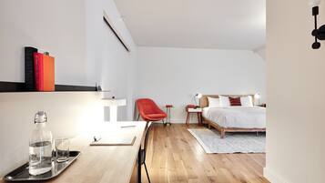 Un dormitorio moderno con suelo de madera, una cama con ropa de cama blanca, una silla roja, una mesita de noche con una lámpara y una estantería con libros.