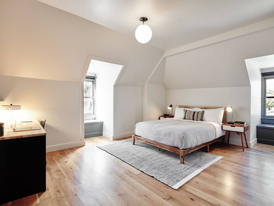 Camera da letto con soffitto a falde, un letto in legno, un comodino con una lampada e un'ampia finestra che lascia entrare la luce naturale.