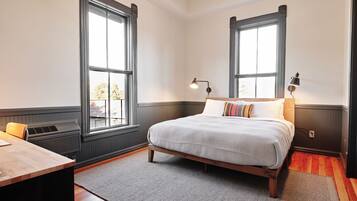 Un dormitorio con cama, dos ventanas, un escritorio y una lámpara.