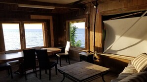 A cozy room with wooden walls and large windows overlooking a body of water. There is a table with chairs, a sofa, and a small table with a lamp.