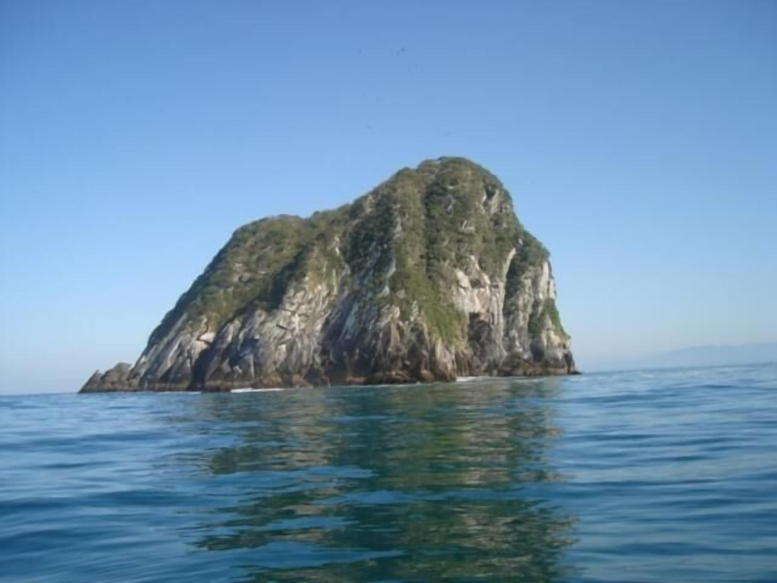 A large, rocky island with steep cliffs and sparse vegetation, surrounded by calm blue waters.