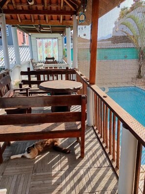 A covered outdoor area with a pool, wooden furniture, and a cat lying on the floor. - Hotel Recanto Do Sol (Cananeia)