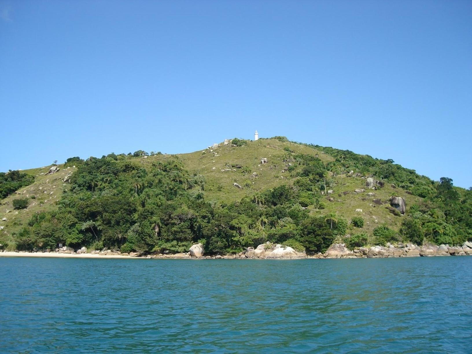 Paisagem costeira com uma colina onde se destaca um farol, rodeada por vegetação densa e um mar calmo.