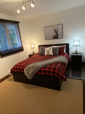 Une chambre à coucher comprenant un lit, deux tables de chevet, une fenêtre avec des stores, un tableau accroché au mur et un luminaire au plafond.