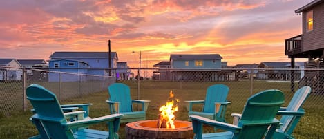 A fire pit with chairs in a grassy area, surrounded by houses and a sunset sky.