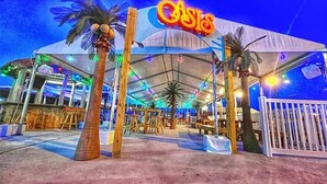 A large, open-air venue with a prominent 'CASIO' sign, featuring a covered seating area with tables and chairs, palm trees, and a white fence.