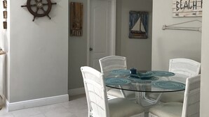 A dining area with a round glass table, white wicker chairs, a ship's wheel decoration, and a 'BEACH' sign on the wall. - Stay in Paradise (Key Largo)