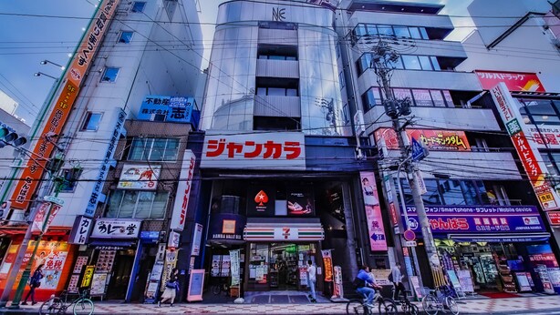 A commercial building with multiple storefronts, including a visible shoe store and a 7-Eleven convenience store. - bnb+ Namba - Hostel (Osaka)