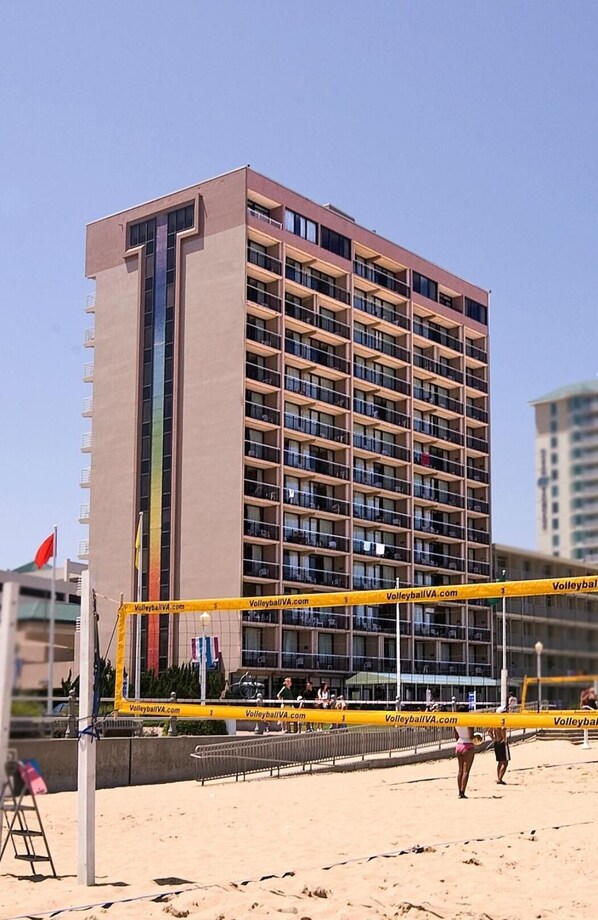 A beach volleyball court with a net and a yellow banner, a tall building in the background, and a flag on the left. - Four Sails Resort - Oceanfront by the Neptune Statue (Virginia Beach)