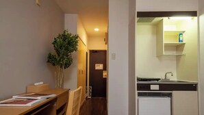 Una cocina compacta con refrigerador, pileta y una pequeña mesada. Hay una zona de comedor con mesa y sillas, y una planta en maceta.