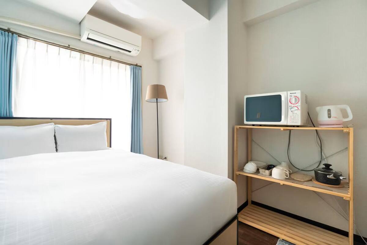 Habitación de hotel con cama, equipo de aire acondicionado, lámpara, estante con electrodomésticos de cocina y ventana con cortinas.