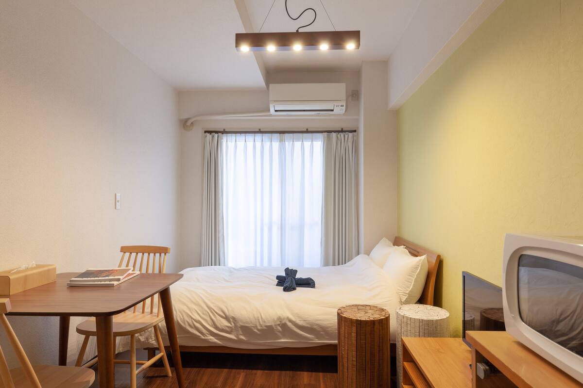 Habitación de hotel pequeña con cama, escritorio, silla, televisión y ventana con cortinas.