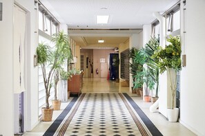 Et hotel med korridor, et mønstret tæppe, plantekasser og en receptionsdisk.
