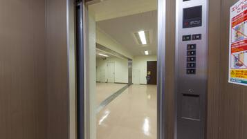An elevator interior with a control panel, emergency exit sign, and a corridor leading to a door.