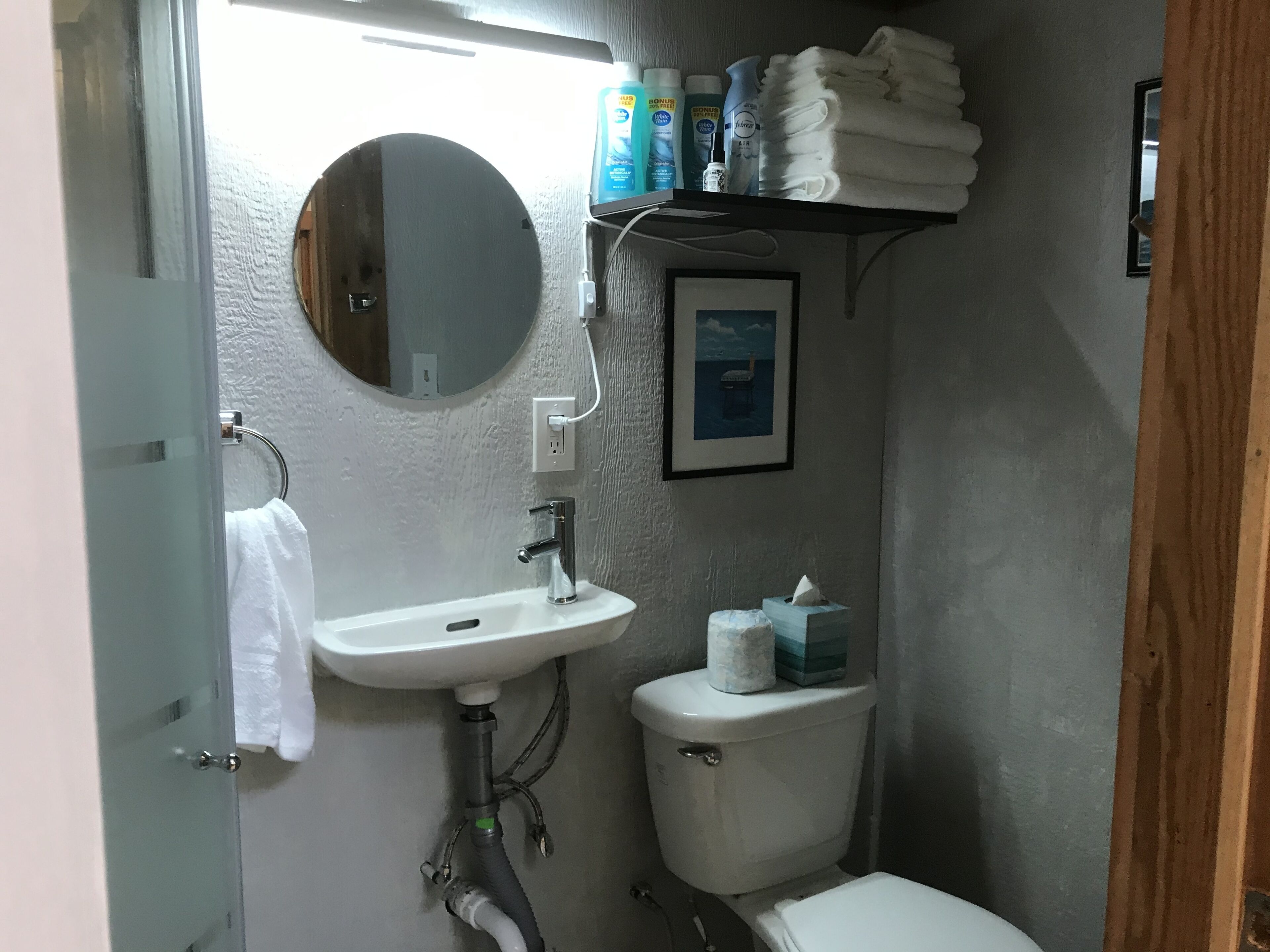Un baño pequeño con un lavamanos blanco, un espejo redondo, un inodoro y un estante con artículos de aseo.